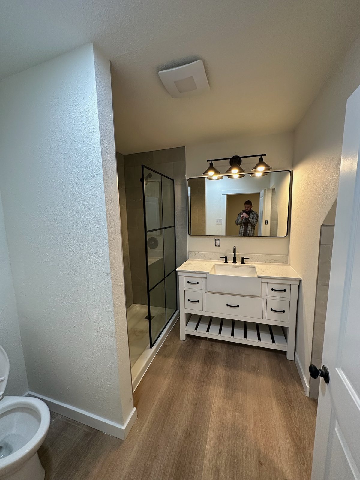 A finished bathroom with a black framed glass walk-in shower, a white farmhouse vanity with black hardware and wood look plank flooring